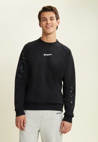 Giovane uomo che sorride, indossando un felpa nera con logo "FREDDY" e pantaloni di felpa grigi chiari, in piedi davanti a uno sfondo semplice e chiaro.