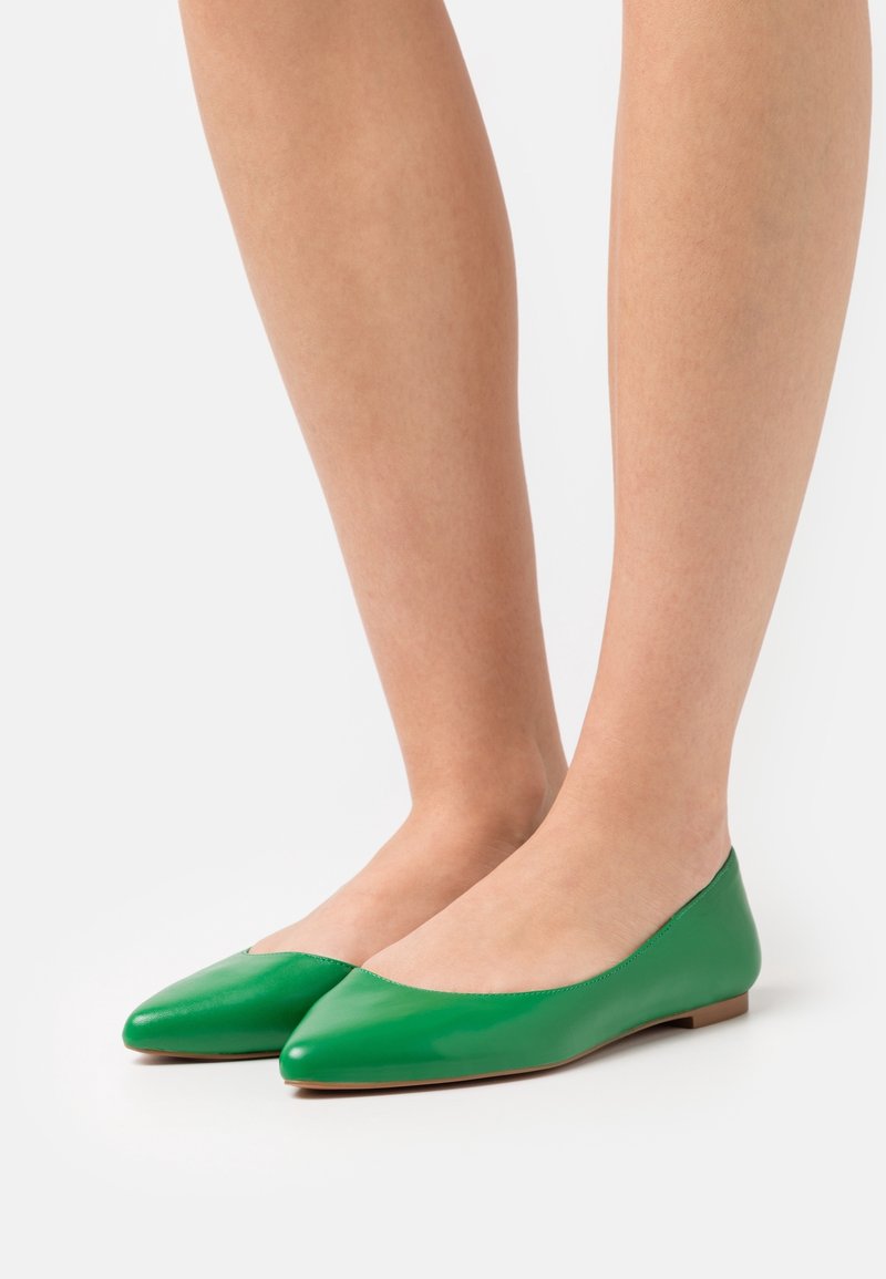 Jambes portant des chaussures plates à bout pointu vert vif sur un fond blanc, montrant une texture de cuir lisse et des semelles basses marron.