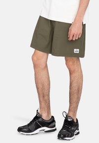 Olijfgroene shorts met een relaxte pasvorm, gestructureerde stof en een merkembleem. In combinatie met zwarte sportieve schoenen met mesh-details.