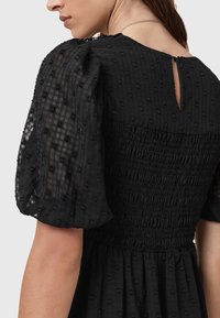 Vestido negro con un cuerpo texturizado y fruncido, y mangas abullonadas. La tela presenta un sutil patrón de cuadrícula y un escote en la espalda con apertura en forma de ojo de cerradura.