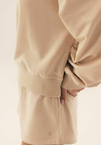 Beżowa bawełniana bluza z kapturem z dżerseju, z prążkowanymi mankietami, w komplecie ze spódnicą w tym samym kolorze. Zauważalny detal z logo na dolnym brzegu obu elementów odzieży.