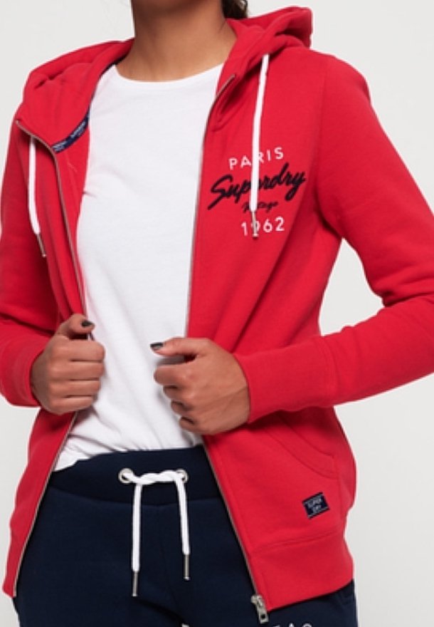 Personne portant un sweat à capuche rouge zippé avec des cordons blancs et un texte noir, un t-shirt blanc et un pantalon bleu marine avec un cordon blanc.