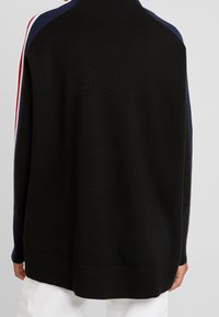 Černý pletený svetr s vysokým výstřihem. Má modré a červené pruhy na ramenech. Jemná textura a žebrované manžety.