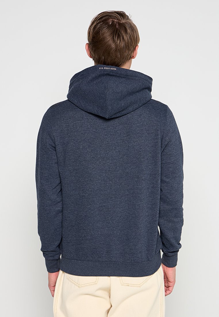 Homme portant un sweat à capuche bleu marine avec "U.S. Polo Assn." sur la capuche, associé à un pantalon beige clair, de dos contre un fond uni.