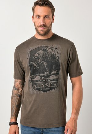 Dospělý muž v hnědém tričku s grafikou burácejícího medvěda chytajícího rybu a textem "Glacier Bay Alaska Od roku 1880."