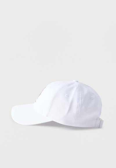 Gorra blanca de béisbol con correa ajustable mostrada desde el lado izquierdo sobre fondo gris.