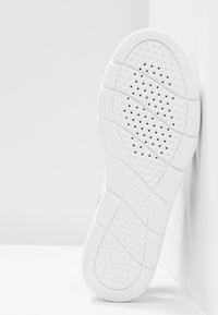 Semelle de chaussure blanche avec grip texturé et petits trous de ventilation, inclinée contre une surface blanche.