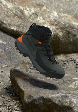 Tmavozelená a čierna turistická topánka s oranžovými akcentmi, označením Gore-Tex a robustnou podrážkou, umiestnená nad skalnatým vonkajším terénom.