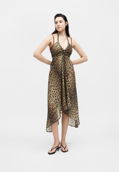 Γυναίκα στέκεται με τα χέρια πίσω από την πλάτη, φορώντας φόρεμα με λεοπάρ ντεσέν και μαύρα σανδάλια με τακούνι, σε λευκό φόντο.