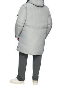 Grijze pufferjas met een capuchon, lange silhoutte en gestikte details. Gecombineerd met een grijze broek en witte sneakers. Achteraanzicht weergegeven.