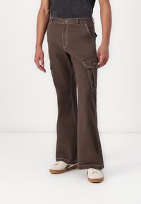 Brown cargo pants with flared legs, contrasting white stitching, multiple pockets, and a relaxed fit. Paired with white sneakers.