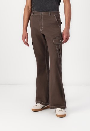Pantalones cargo marrones con piernas acampanadas, costuras blancas en contraste, múltiples bolsillos y un ajuste relajado. Combinados con zapatillas blancas.