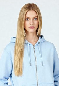 Felpa con zip di colore blu chiaro, con cappuccio regolabile tramite cordino, dalla texture morbida e con dettagli in metallo argentato. Design semplice e tasca a marsupio frontale.