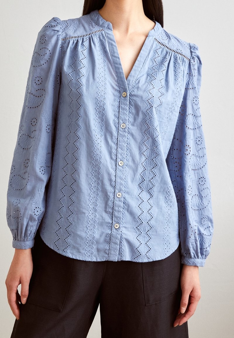 Blouse bleu clair à manches longues avec boutonnière sur le devant, motifs en dentelle ajourée, découpes en zigzag et florales, portée avec un pantalon foncé.