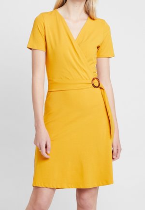 Mujer con un vestido cruzado de manga corta de color mostaza con un cinturón de anillo carey en la cintura, de pie frente a un fondo liso.