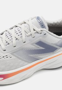 Baskets de sport gris clair avec un dessus en mesh, accents bleus, une bande orange sur une semelle blanche, et un logo bien visible sur le côté et la languette.