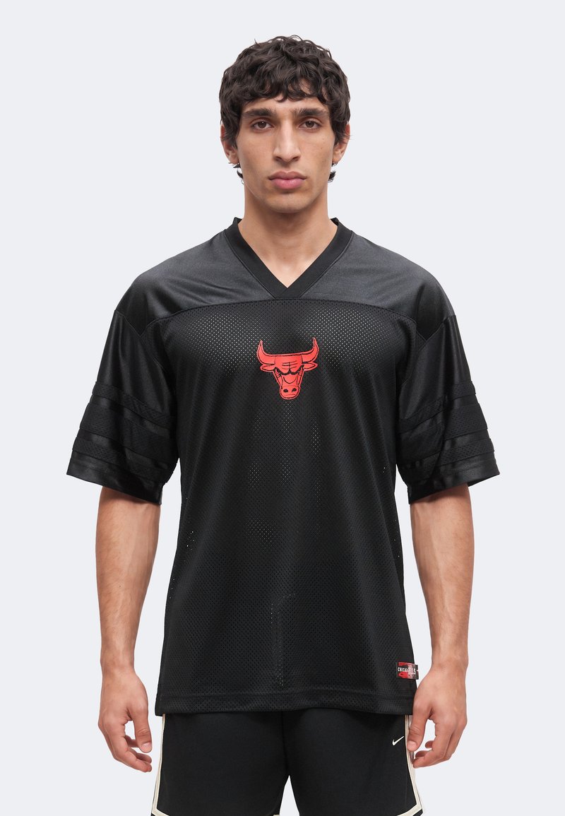 Άνδρας που φοράει μαύρο δίχτυ Chicago Bulls με κόκκινο λογότυπο ταύρου και μαύρο αθλητικό σορτς, στέκεται μπροστά από απλό φόντο.