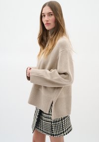 Beige stickad tröja med hög ribbad krage, drop shoulders och ribbad fåll. Kombinerad med en mönstrad svartvit kjol.
