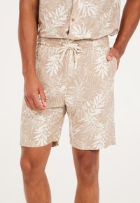 Pantalones cortos beige con un patrón de hojas tropicales en blanco. Fabricados con material ligero, cuentan con una cinturilla elástica y cierre de cordón.