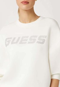 Felpa bianca con scollatura rotonda e maniche larghe. La parte frontale presenta un logo "GUESS" scintillante realizzato in strass.