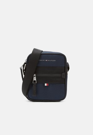 Kompaktní tmavě modrá crossbody taška s černým nastavitelným popruhem, přední kapsou na zip a logem Tommy Hilfiger na strukturované látce.