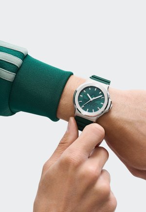 Žalias laikrodis su nerūdijančio plieno korpusu ir tekstūruota žalia audinio apyranke. Ciferblate yra sidabriniai valandų žymekliai ir rodyklės su logotipu.