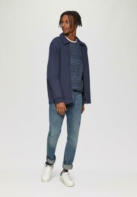 Marineblauer Reißverschlussjäckchen über einem blauen gemusterten Pullover, kombiniert mit hellblauen Jeans und weißen Sneakern. Glatte Texturen und lässiges Design.