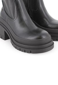 Botas de cuero negro hasta el tobillo con punta redondeada, suela de goma estriada y tacón grueso; cuentan con una cremallera lateral para ponerlas y quitarlas fácilmente.