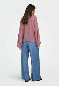 Kvinde med langt brunt hår iført en løs mauve bluse med små prikker og ballonærmer, kombineret med bredbenede lyseblå bukser.