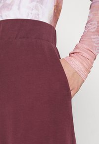 Bordeaux rok met een soepele stof, voorzien van een tailleband en zijzak, gecombineerd met een nauwsluitend, gestructureerd langemouwen shirt in pastelroze.