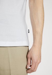 Mężczyzna ubrany w beżowe spodnie i białą koszulę z małą czarną etykietą "BOSS" na dolnej krawędzi koszuli, z ręką swobodnie opuszczoną wzdłuż ciała.