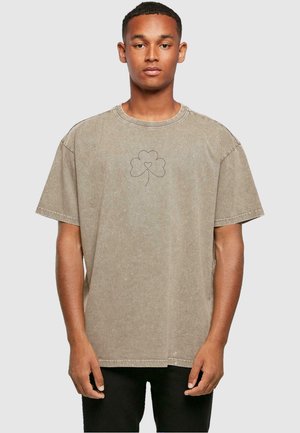 Beige oversized t-skjorte laget av myk bomull. Har et lite svart kløverdesign på brystet og synlige sømmer. Korte ermer.
