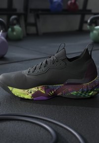 Chaussure de sport noire avec semelle géométrique multicolore posée sur le sol de la salle de sport avec kettlebells et équipement d'exercice en arrière-plan.