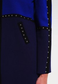 Blau-schwarze Strickjacke mit silbernen Nieten an den Nähten und Taschenrändern, ausgestattet mit kontrastierenden geometrischen Mustern und strukturiertem Stoff.