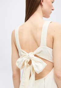 Γυναίκα που φοράει κρεμ αμάνικο φόρεμα με μεγάλο δεμένο φιόγκο στην ανοιχτή πλάτη, που δείχνει τους ιμάντες των ώμων και μια πλαϊνή διάτρηση.