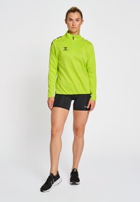 Heldergroen lange mouw sporttop met een kwartrits en zwarte schouderaccenten, gecombineerd met zwarte shorts en zwarte sportschoenen.