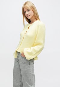 Rumeno pleteno jopico z okroglim izrezom, rebrasto teksturo in zapiranjem na gumbe. Ima nablaščen rokav in sproščen kroj.