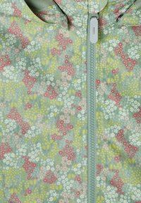 Veste zippée à motifs floraux avec un fond vert pastel, ornée de petits motifs floraux roses, jaunes et blancs ; texture de tissu lisse.