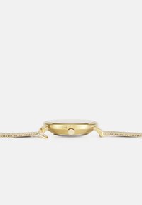 Montre dorée avec un boîtier circulaire, bracelet en maille mince et couronne proéminente. Surface en verre lisse, design minimaliste et finition brossée.
