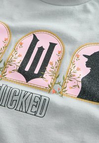 Sivá bavlnená tričko so grafickým dizajnom, s ružovými oblúkmi, kvetinovými akcentmi, výrazným čiernym textom "WICKED" a trblietavými detailmi.