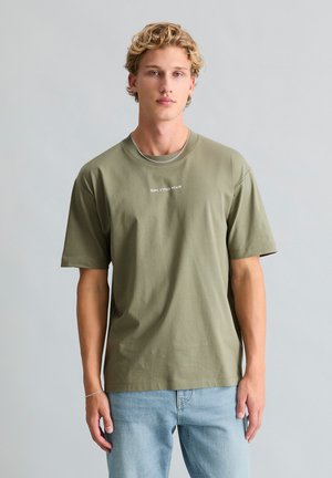 Blondyn z kręconymi włosami nosi oliwkową zieloną koszulkę z napisem „Marc O'Polo Denim” oraz jasnobłękitne dżinsy, stoi przy jednolitej szarej ścianie.