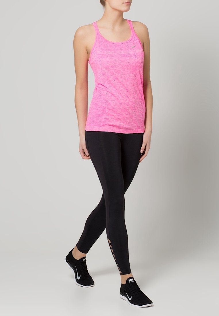 Femme portant un haut de sport sans manches rose, des leggings noirs avec des détails en mesh et des baskets noires Nike, debout, jambes croisées.