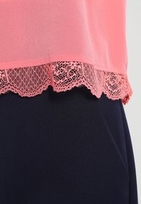 Haut en chiffon rose avec un ourlet en dentelle ondulé, associé à un tissu uni bleu marine. La dentelle présente des motifs complexes le long du bord.
