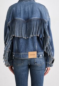 Veste en jean avec de longues franges sur le dos et les manches, portée avec un jean assorti, étiquetée "HUGO" dans le bas du dos.