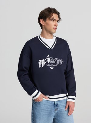 Giovane uomo che indossa un maglione blu navy a scollo a V con strisce bianche e testo grafico, abbinato a jeans azzurri, in piedi davanti a uno sfondo neutro.