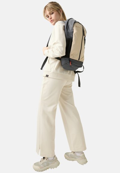 Backpack in gray and beige fabric, featuring a zip closure and red accent, worn over a beige sweatshirt and wide-leg pants.