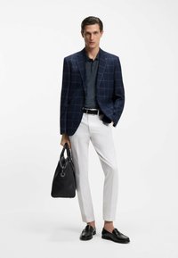 Granatowa marynarka w kratę na ciemnym polo, białe spodnie, czarne mokasyny i czarna skórzana torba. Gładkie tekstury i dopasowany krój.