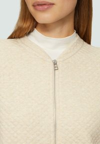 Beige gewatteerde ritssluitingjas met een hoge hals en subtiele textuur. Heeft een voorrits en een gladde binnenlaag.