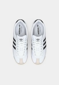 Bijele Adidas tenisice s tri crne pruge, bež prednjim dijelom i bijelim vezicama. Teksturirani gornji dio s logotipom na jeziku.