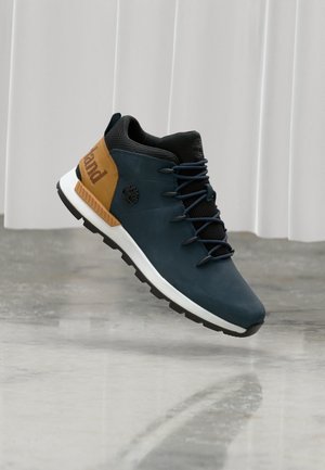 Navyblauwe en beige Timberland sneaker met zwarte veters, witte zool en logo, zwevend boven een gepolijste betonnen vloer.